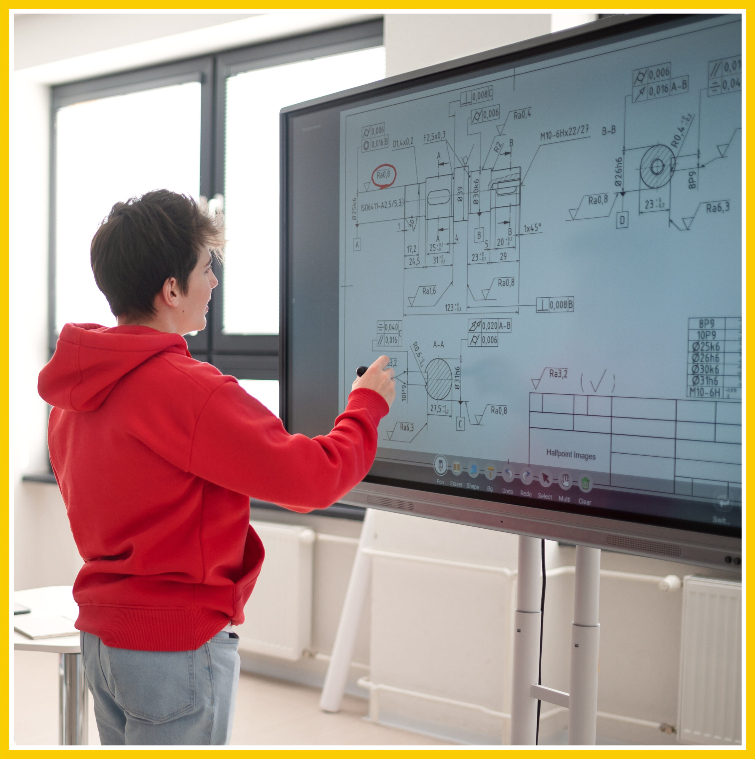 A high-school or college student standing to the left in the image, writing the answer to an equation or drawing out measurements for something on an interactive display screen.