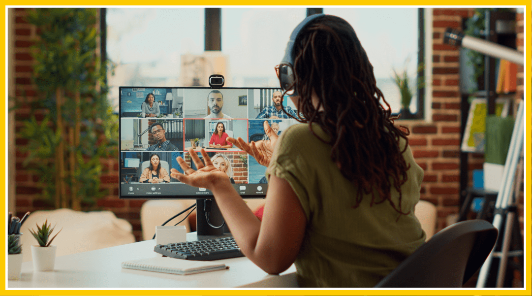 View of a woman sitting at her desk at home, facing away from the camera. There are various others on the computer screen in front of her. She holds her hands up as she talks with the group remotely.