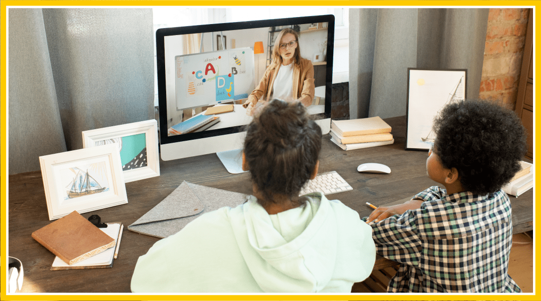 Two young children on a video call lesson with their teacher. They are facing away from the camera while their computer screen faces us.