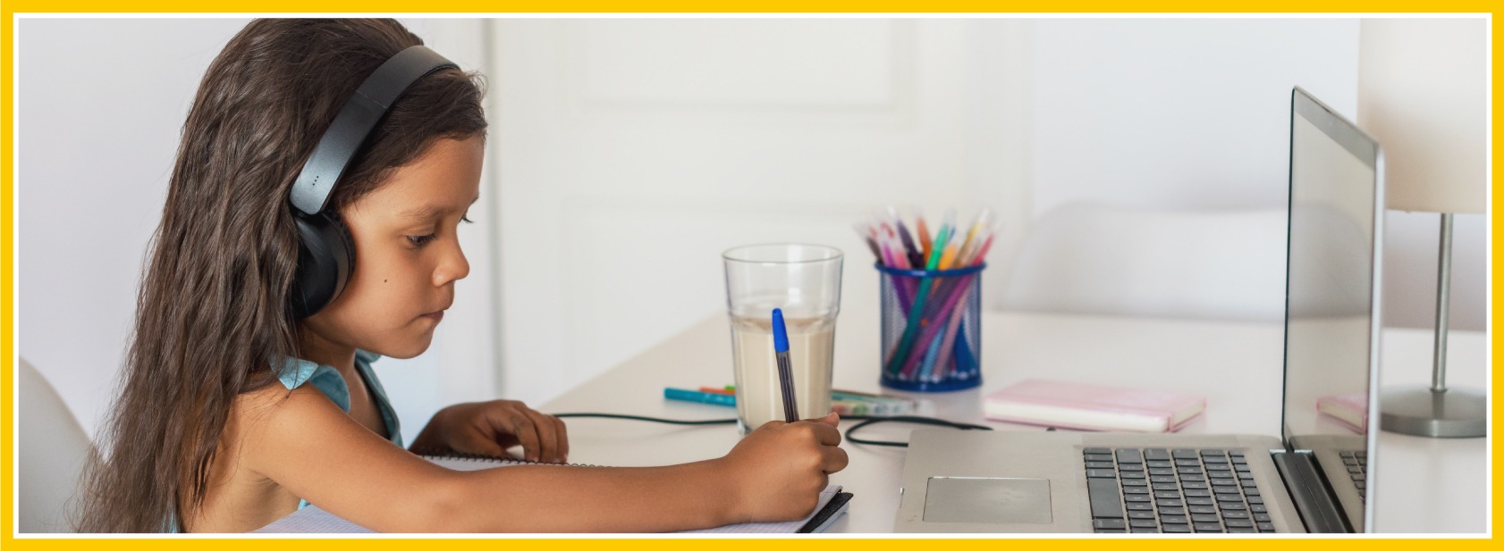 Side view of a young girl taking notes while she sits at a desk with her laptop in front of her.