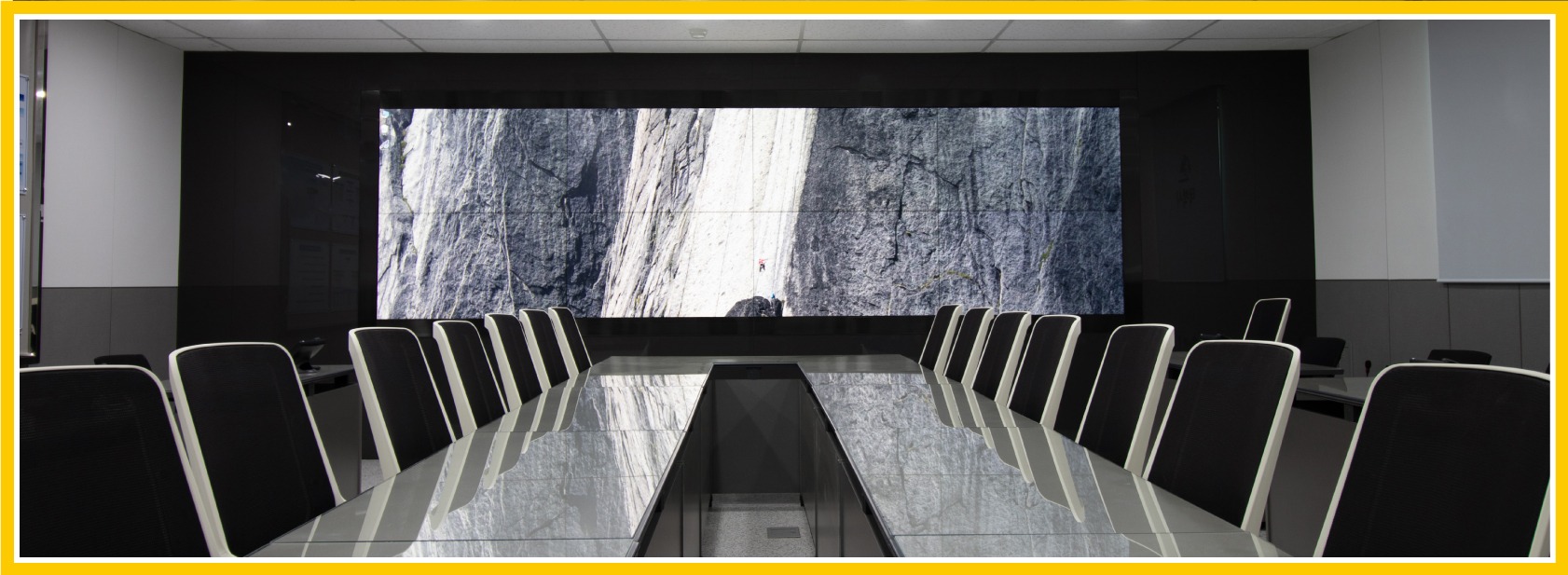 Framed image of video walls in a business setting: A view of an empty conference table from the surface of the table, looking off towards a row of screens in the background. They're all on and linked together.