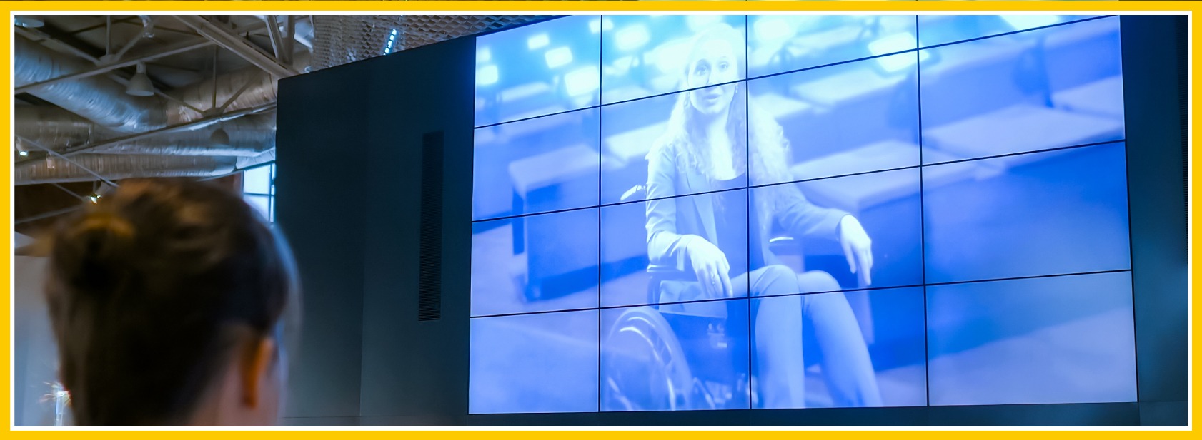 Framed image of a video wall in a learning environment setting: A close-up of a woman from the back as she watches a row of screens all working together to show a woman in a wheel chair at a much larger scale.