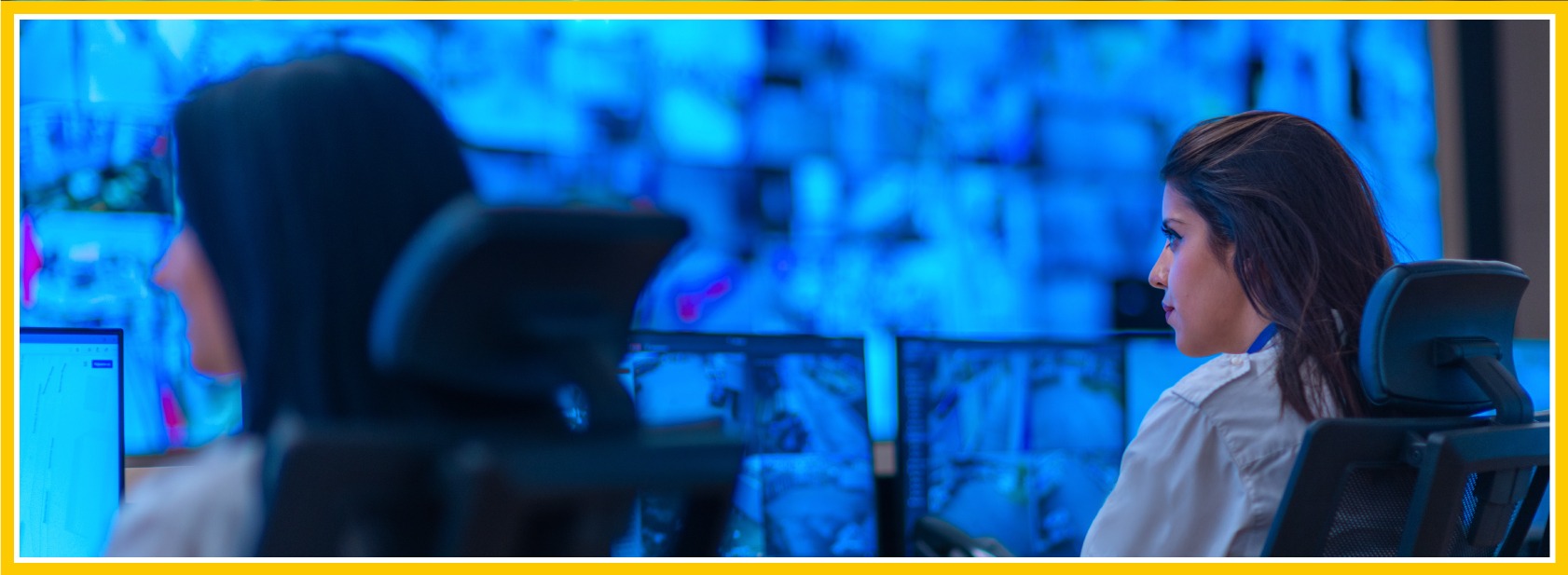 Framed image of video walls in a security setting: Two women are pictured in the foreground sitting at computers with various locations on-screen. In the background is a much larger wall of video feeds from different locations around the building. The woman on the right is looking up at this large wall and monitoring various screens.