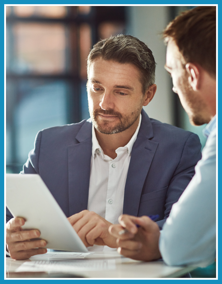 Two men collaborating together; focus is on one man with the other men just barely off to the right facing away from the camera. The first man is looking down at a tablet in his hand.