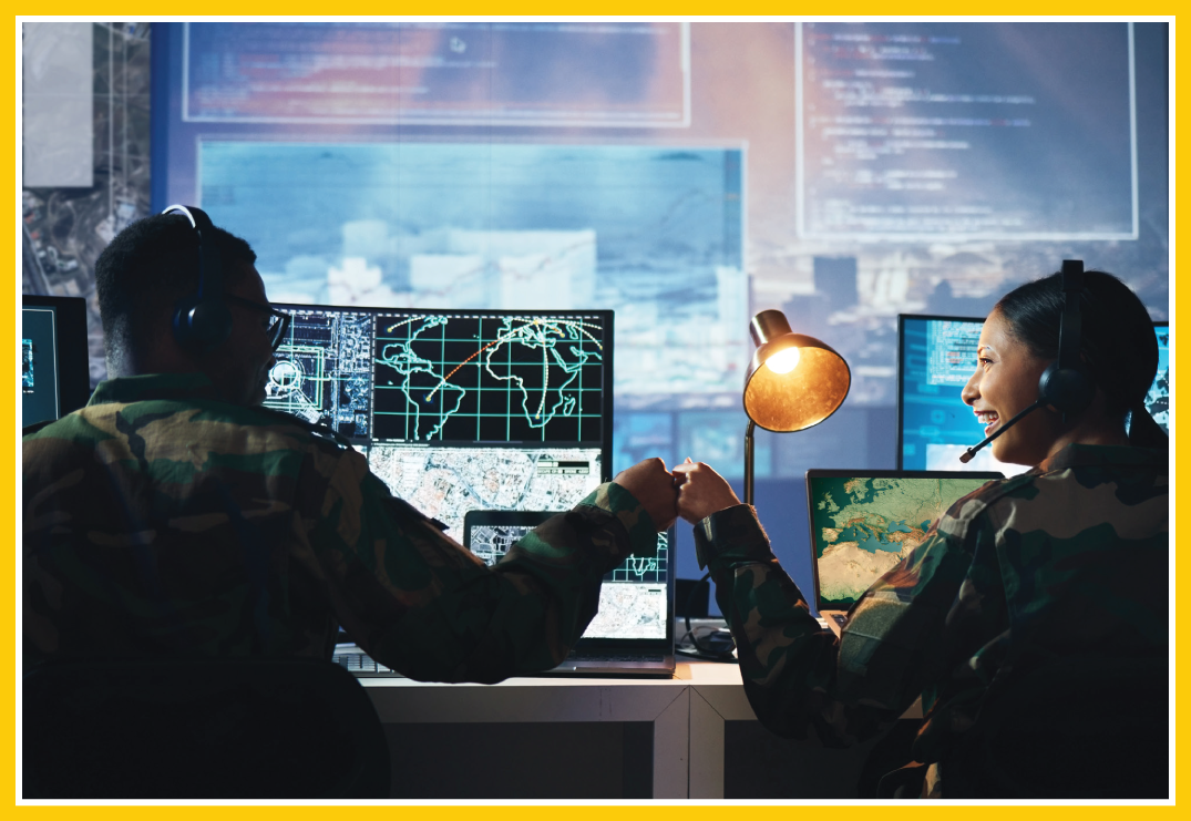 Two military officials fist bumping in front of various screens, including a large video wall in the background.