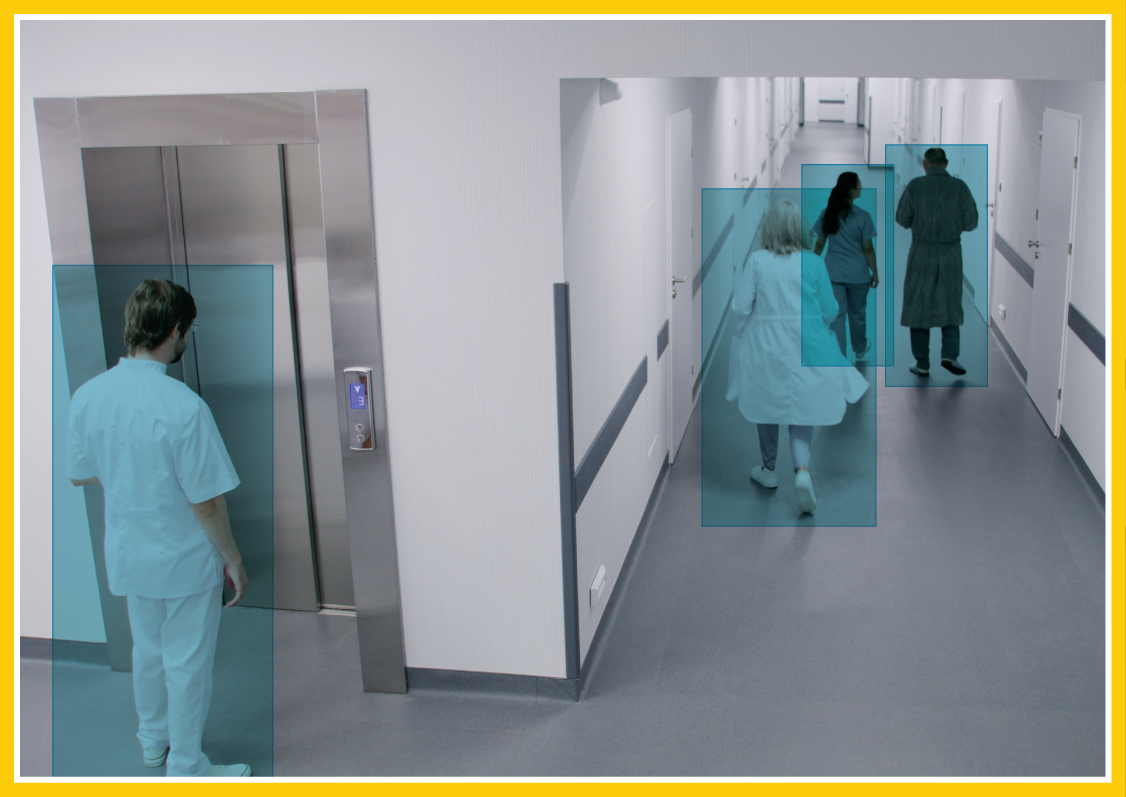 Image from the perspective of a surveillance camera; people are walking down a hallway in a healthcare facility and are highlighted via AI-powered software.