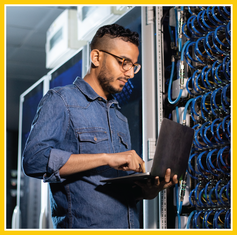 A man is standing in front of an infrastructure unit full of various cables. He's looking down at a laptop balanced in his hands and typing up a note.