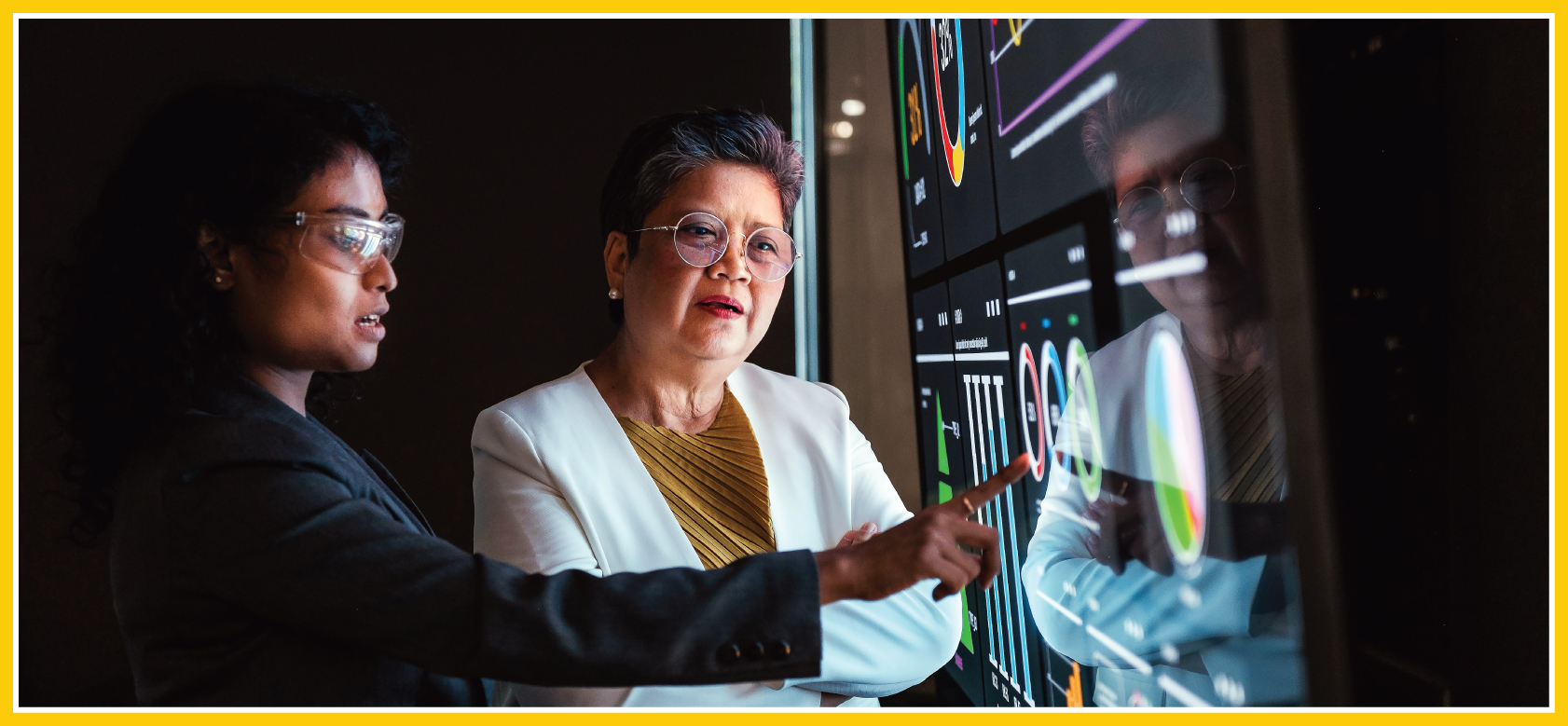 Two decisionmakers are working together in front of a large interactive display, using it as their production dashboard as both women discuss the logistics and details