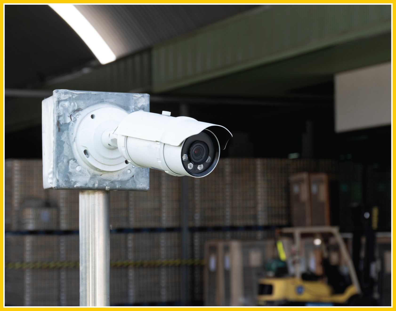 Close-up of a security camera observing a manufacturing warehouse. Rows of pallets and crates are in the background with a forklift off to the right.