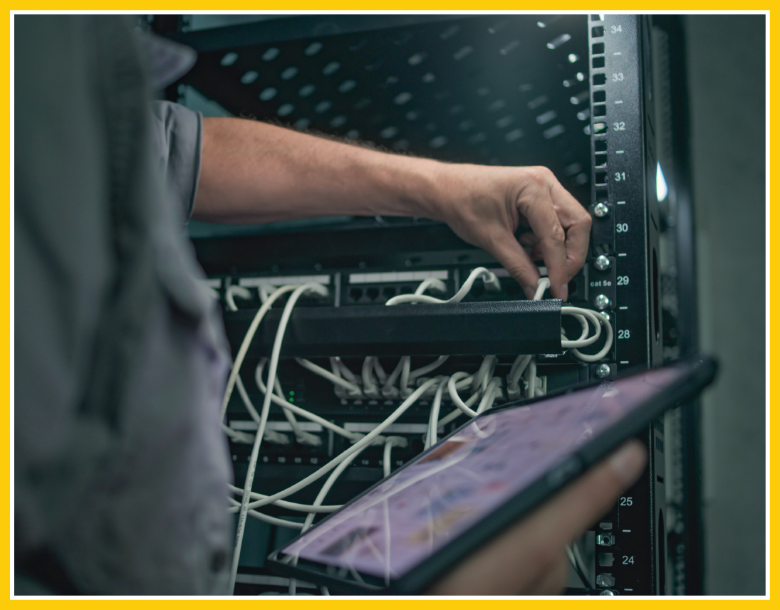 Close-up of a person setting up network infrastructure. They're plugging in different cables with their left hand while referring back to a tablet with a guide in their right hand.
